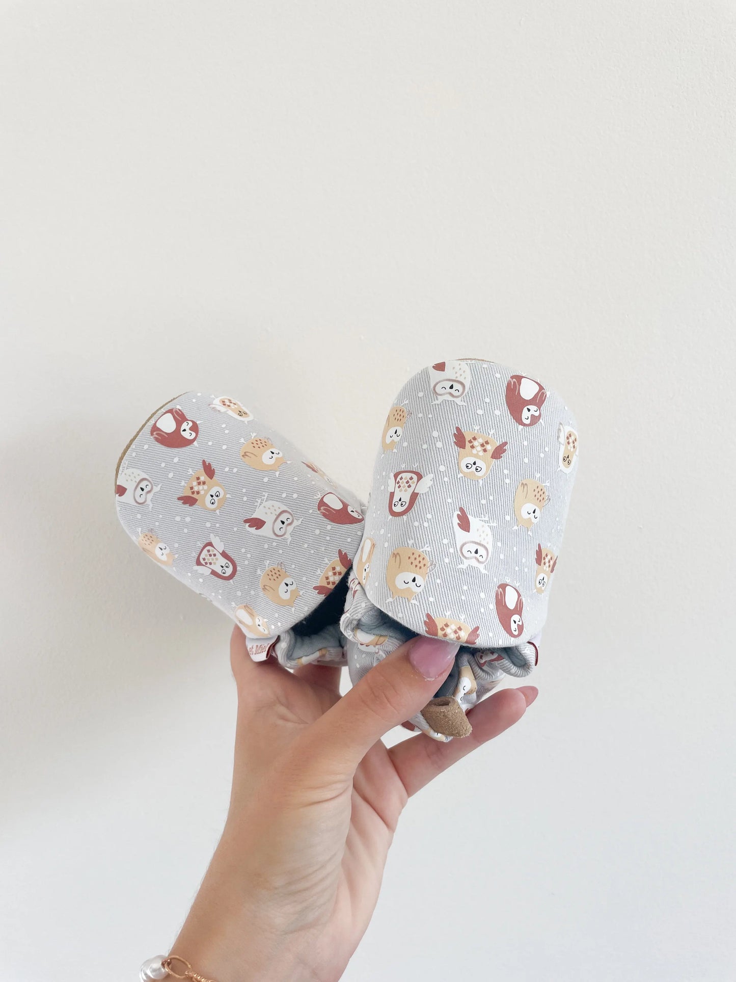 Chaussons souples bébé avec motifs hiboux colorés sur fond gris clair, tenus en main sur fond blanc