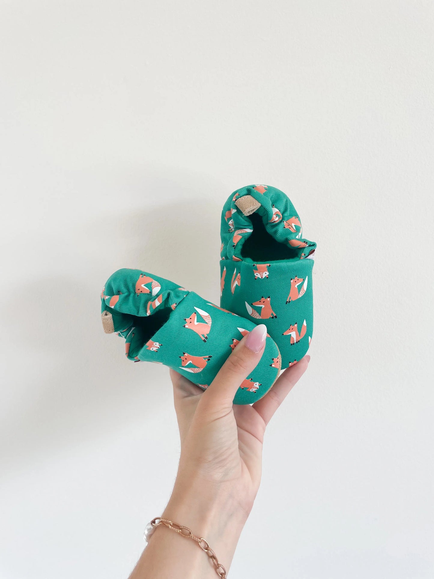 Chaussons souples bébé avec imprimé renards sur fond vert sapin, tenus en main sur fond blanc.