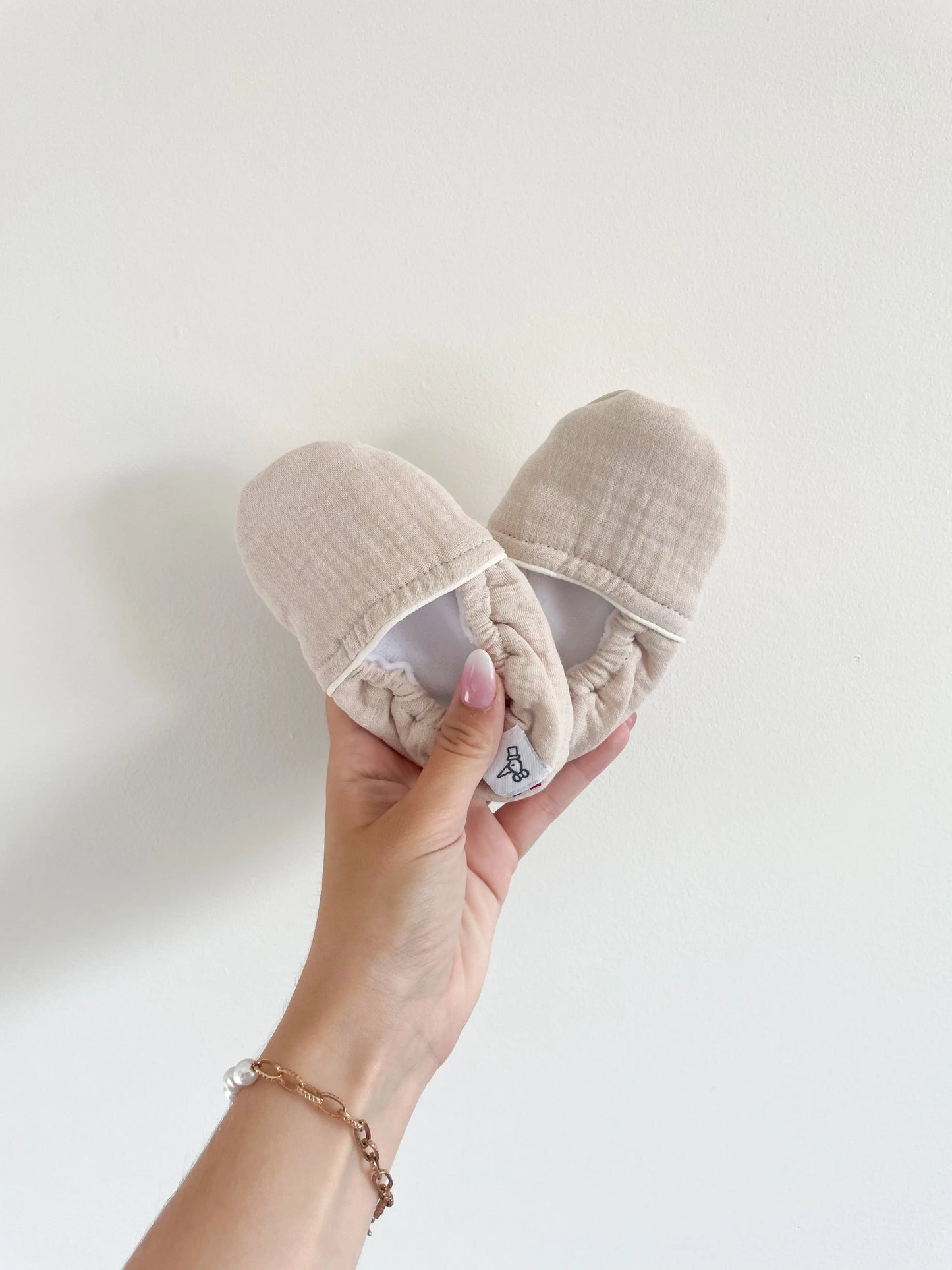 Chaussons souples bébé coloris sable, semelle antidérapante et tissu doux, tenus à la main