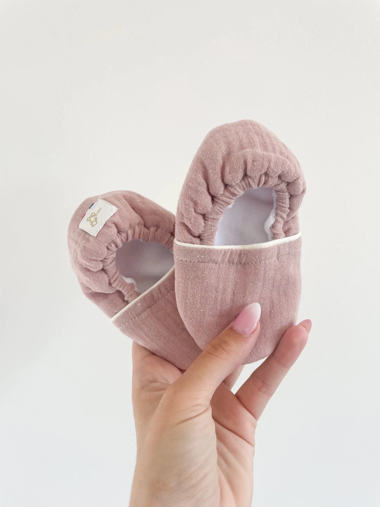 Chaussons souples bébé vieux rose en cuir grainé, tenus à la main sur fond blanc avec étiquette little cigogne, semelle antidérapante