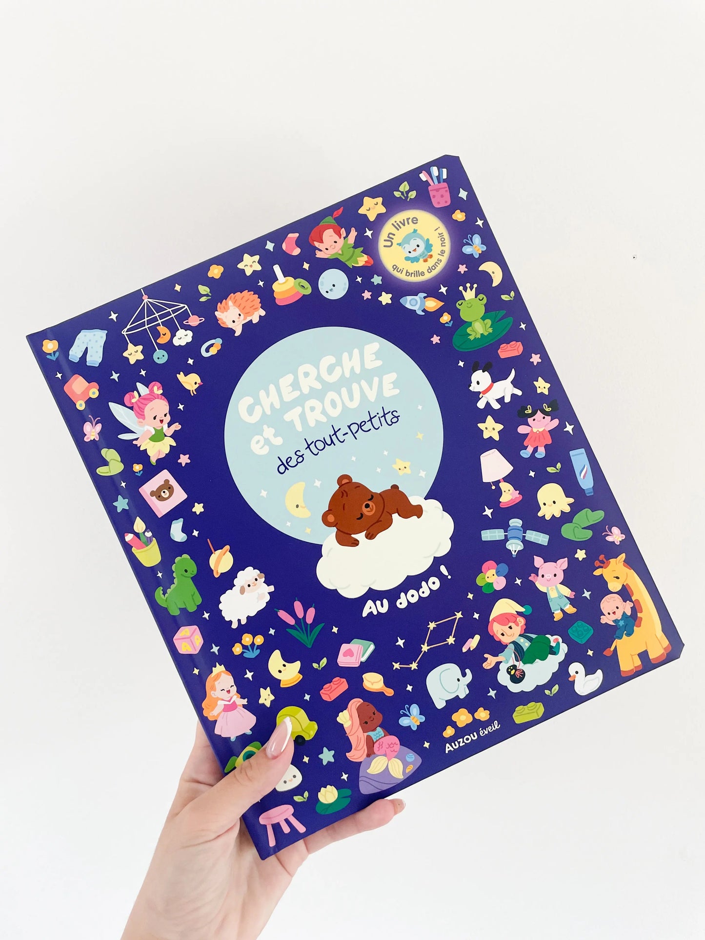 Livre Cherche et trouve des tout petits - Au dodo dès 1 an. Page de couverture avec des details. Livre tenu dans une main sur un fond blanc.
