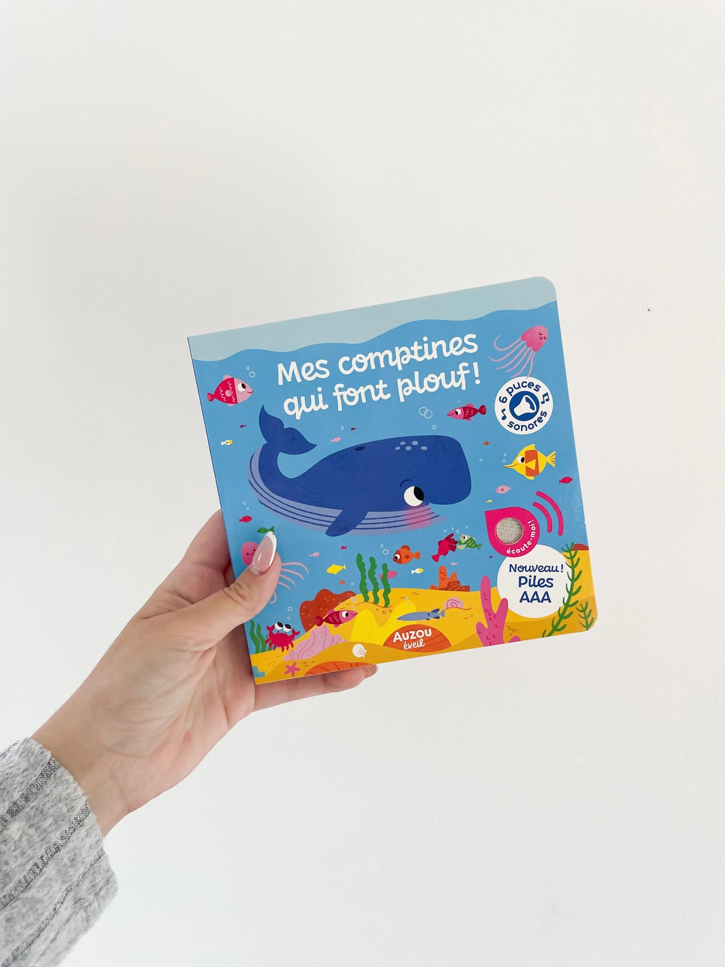 Livre sonore - Mes comptines qui font plouf dès 1an. Sur le thème de l'eau. Page de couverture du livre musicale avec baleine. Livre dans une main sur fond blanc