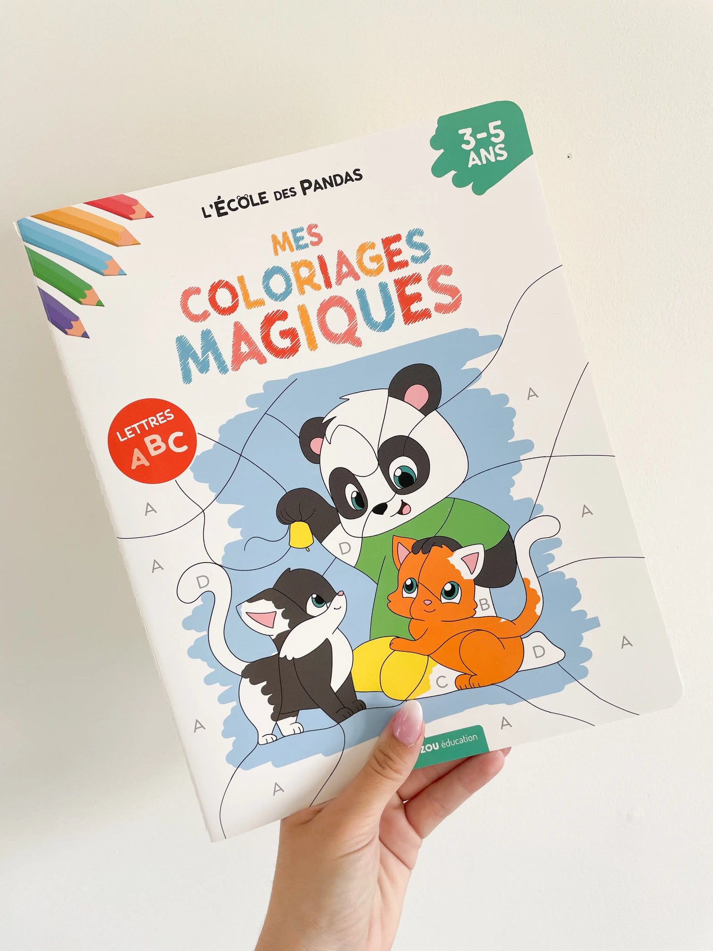 Coloriages magiques sur les lettres thème panda pour les 3-5 ans. Page de couverture. L'école des panda. Livre dans une main sur un fond blanc.