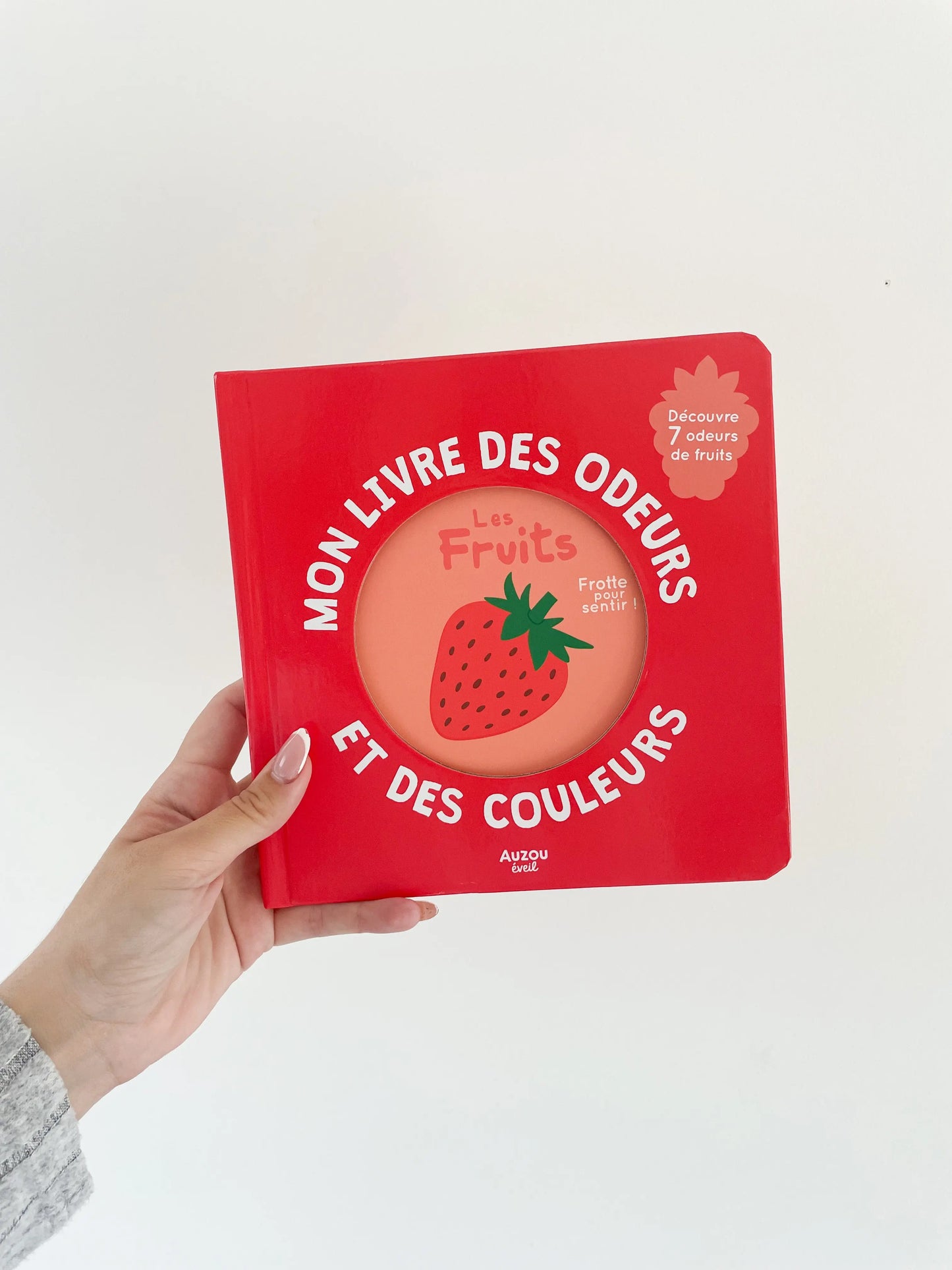 Mon livre des odeurs et des couleurs - Sur les fruits. Page de couverture du livre de couleur rouge avec une fraise. Dès 1an. Livre dans une main sur fond blanc