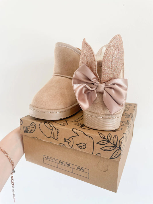 Paire de bottines beige pailletées pour bébé fille avec oreilles de lapin, posées sur une boîte en carton