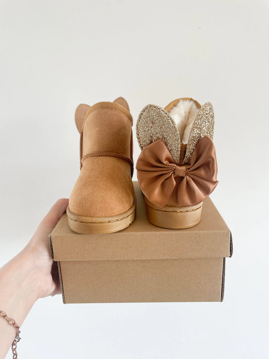 Paire de bottines camel pour bébé avec oreilles de lapin et nœud, posées sur une boîte en carton 
