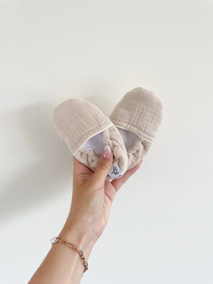 Chaussons souples bébé coloris sable, semelle antidérapante et tissu doux, tenus à la main