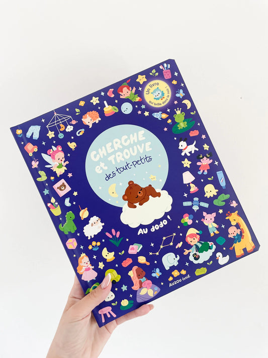 Livre Cherche et trouve des tout petits - Au dodo dès 1 an. Page de couverture avec des details. Livre tenu dans une main sur un fond blanc.