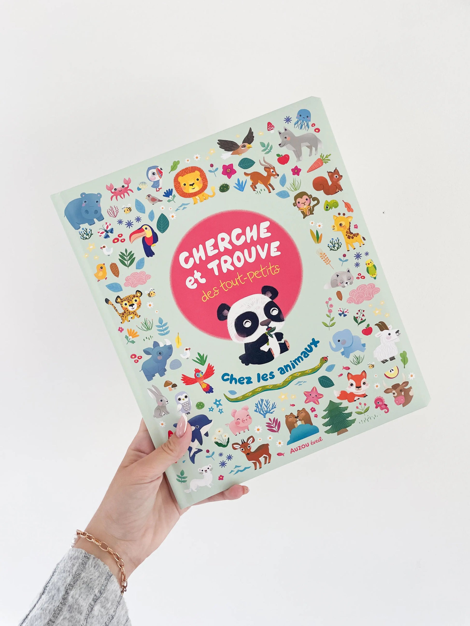 Livre cherche et trouve des tout petits - Chez les animaux. Page de couverture avec un panda et des petit élements. Livre dans une main sur un fond blanc.