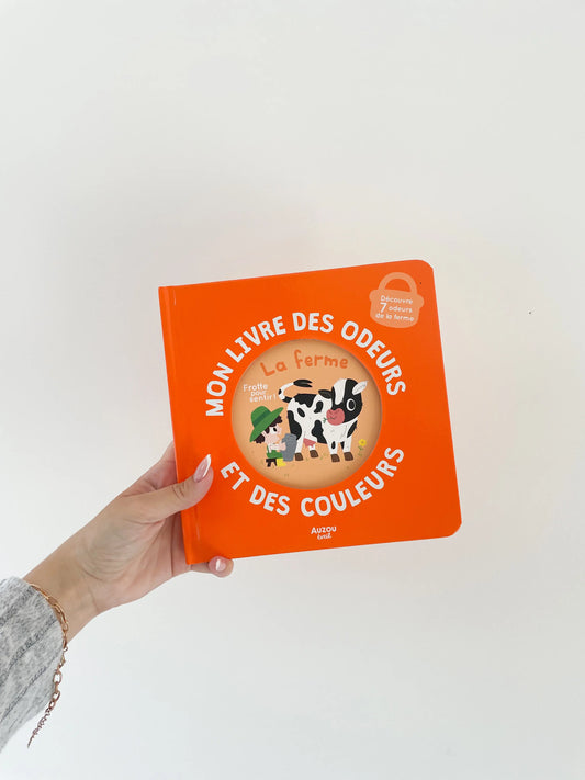 Mon livre des odeurs et des couleurs - Thème de la ferme, vache. Couverture du livre orange. Livre des odeurs dans la main sur fond blanc
