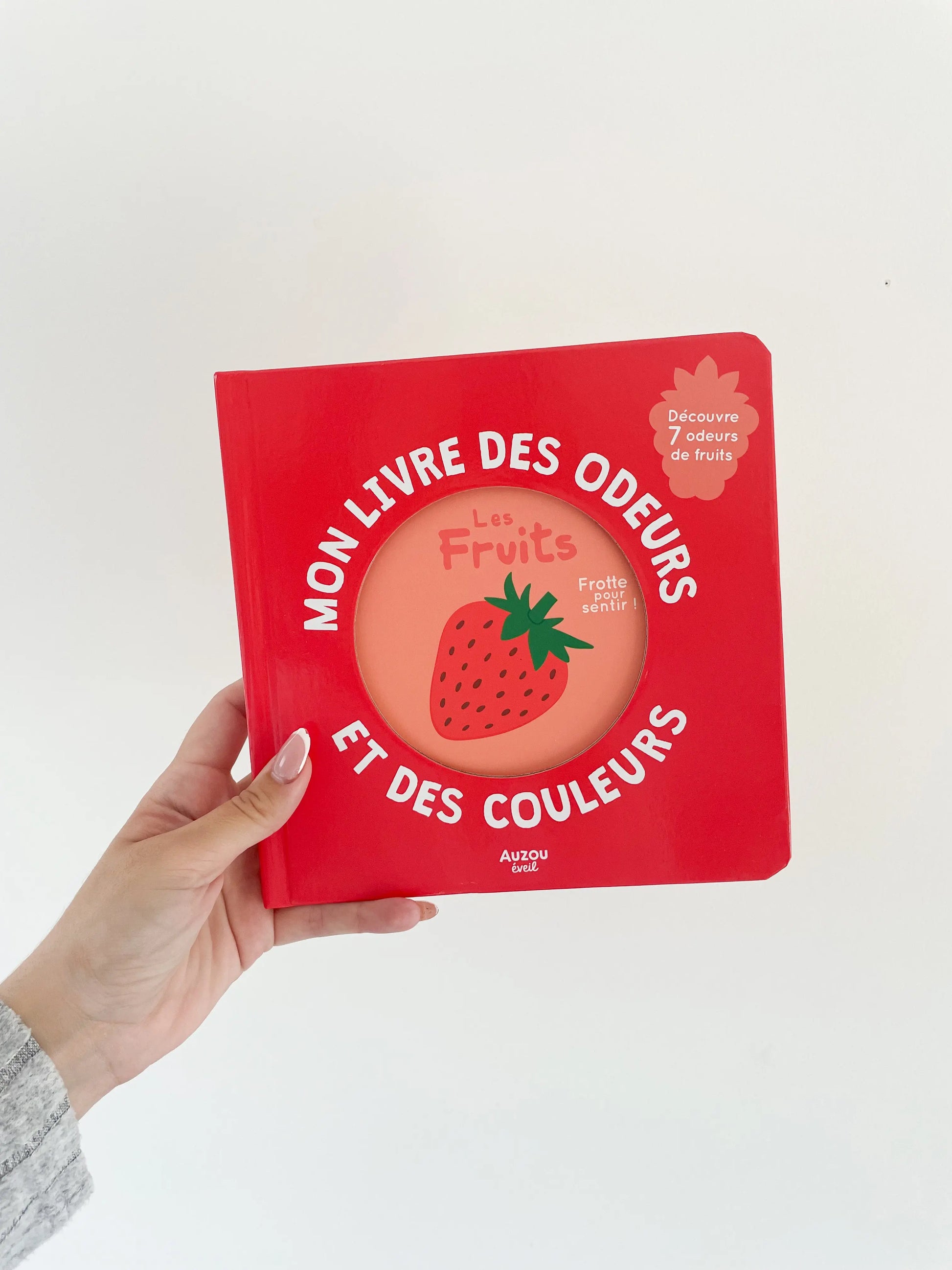 Mon livre des odeurs et des couleurs - Sur les fruits. Page de couverture du livre de couleur rouge avec une fraise. Dès 1an. Livre dans une main sur fond blanc