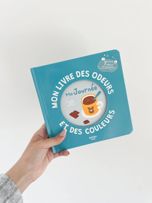 Mon livre des odeurs et des couleurs - Ma journée. Page de couverture, livre a sentir. Livre dans une main sur fond blanc.